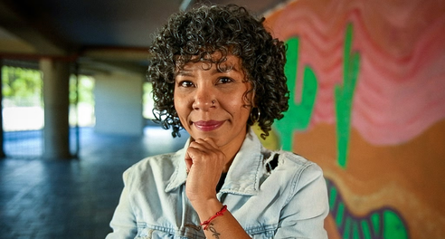 A woman smiles to camera, standing in front of a wall painted with a mural of cactuses