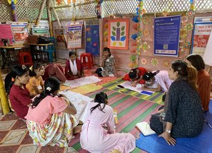 A group of about 10 young girls sit on the floor in a circle with two female facilitators