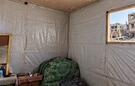 Interior of a makeshift room lined with plastic sheeting, with a chair, small table and personal belongings. Through an open window, heavily damaged buildings and rubble are visible outside.
