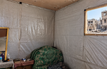 Interior of a makeshift room lined with plastic sheeting, with a chair, small table and personal belongings. Through an open window, heavily damaged buildings and rubble are visible outside.