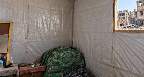 Interior of a makeshift room lined with plastic sheeting, with a chair, small table and personal belongings. Through an open window, heavily damaged buildings and rubble are visible outside.
