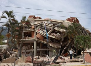 A 3 or 4 story building that has partially collapsed. Two people face the building surveying the damage.