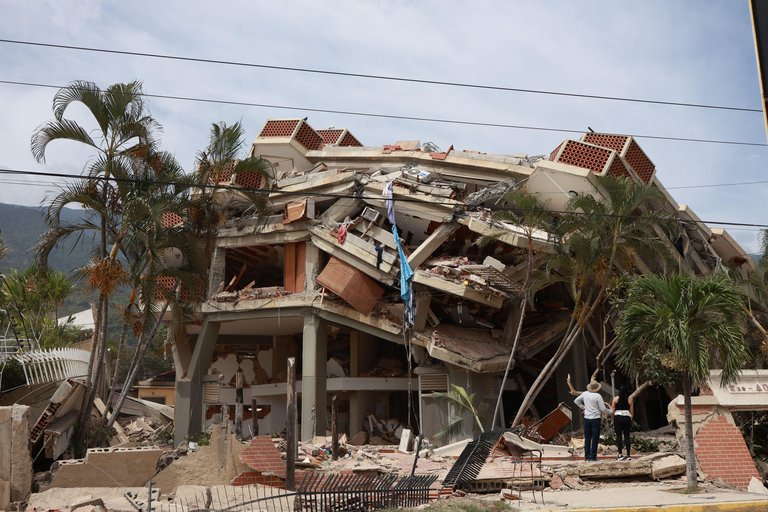 A 3 or 4 story building that has partially collapsed. Two people face the building surveying the damage.
