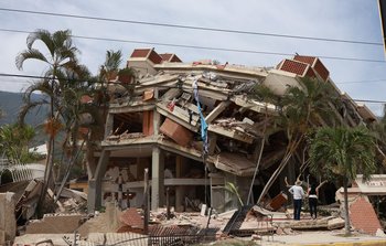 A 3 or 4 story building that has partially collapsed. Two people face the building surveying the damage.
