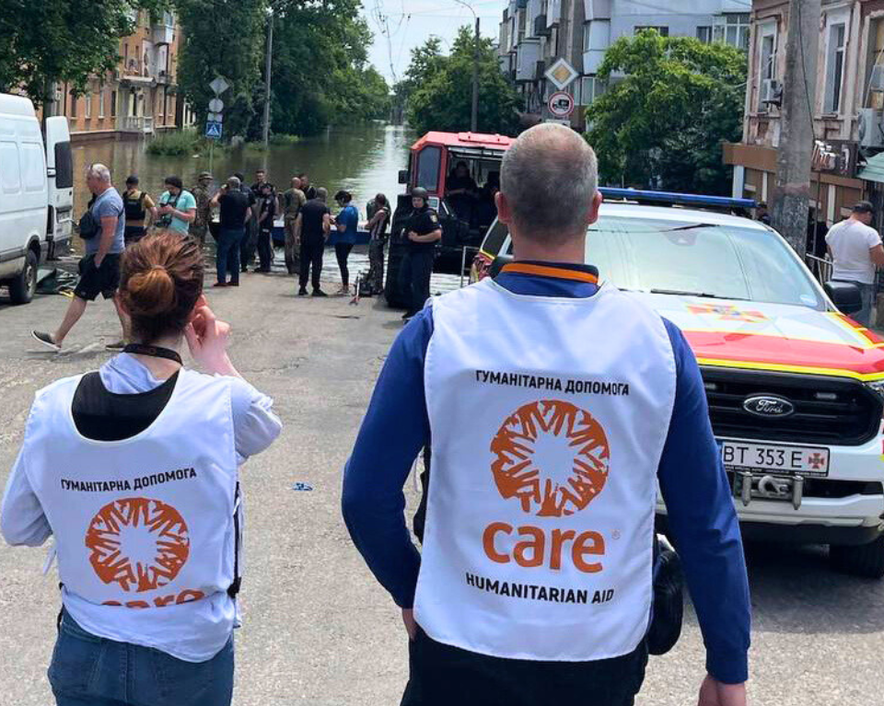 The backs of two aid workers wearing CARE vests walking towards a group of people standing by a canal