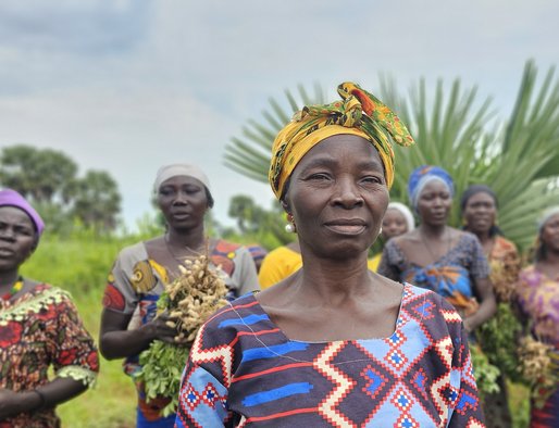 A woman looks determinedly to camera, standing in a field in the foreground in front of a group of people who are holding crops