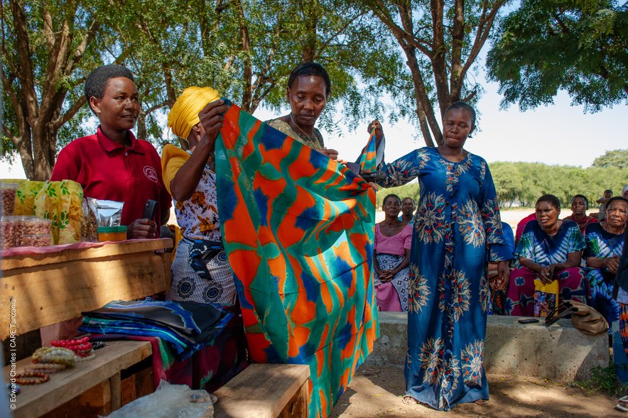 A group of four women in brightly coloured clothes holding up some African textiles. Several other women look on in the background.