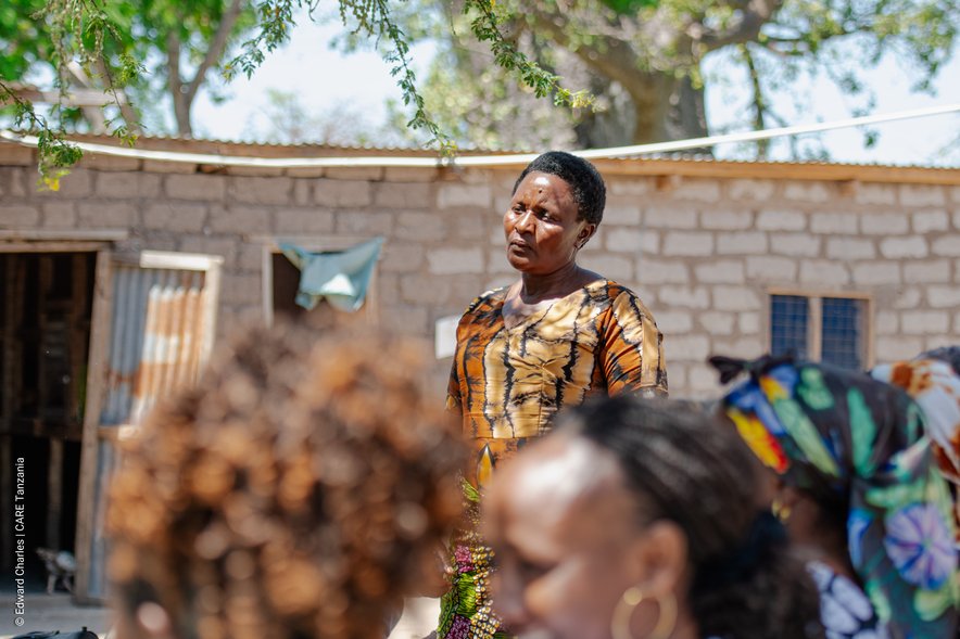 A woman speaking publicly to a group of people, a few of whom are visible in the foreground