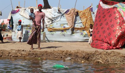 A woman holds a baby in front of a makeshift shelter. Flood waters are in the foreground.