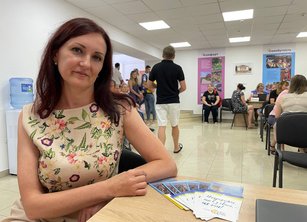 A woman sits at a desk in a community centre. Several people are visible in the background - some at desks, some having a group discussion.