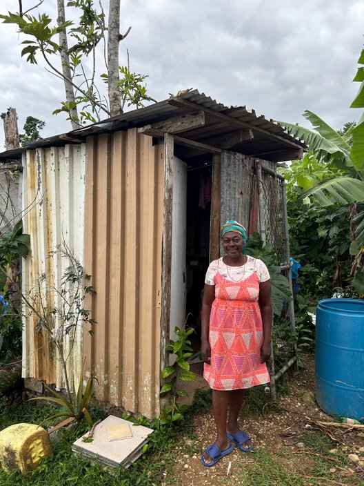 A woman stands next to an outhouse building