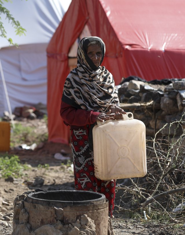 A woman stands next to a well holding a plastic water container. Tents are visible in the background.