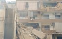 A block of flats partially destroyed by bombing. A woman looks out from a top floor balcony.
