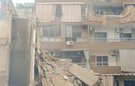A block of flats partially destroyed by bombing. A woman looks out from a top floor balcony.