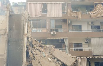 A block of flats partially destroyed by bombing. A woman looks out from a top floor balcony.