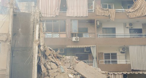 A block of flats partially destroyed by bombing. A woman looks out from a top floor balcony.
