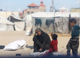 A woman sits with two children outside a tent with her hand to her head in despair