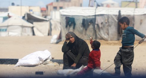 A woman sits with two children outside a tent with her hand to her head in despair