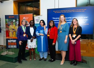 A group of seven women speakers stand together in front of banners at an event