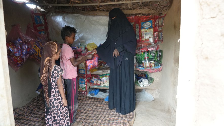 A woman sells items to two young children in a simple grocery store