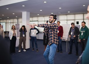 Omer is holding a microphone, speaking at a Shelter workshop. Other workshop participants stand in a circle behind him.