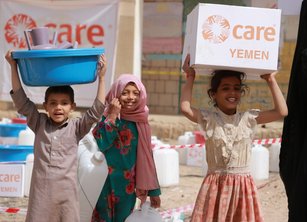 Three children carrying various supplies including a large box with the CARE Yemen logo