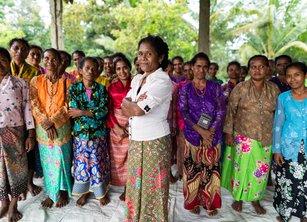 A group of about 20 women stand with their arms folded looking to camera; a woman in a white shirt stands in the foreground