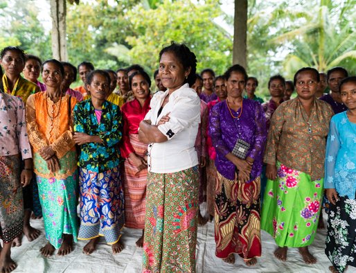 A group of about 20 women stand with their arms folded looking to camera; a woman in a white shirt stands in the foreground