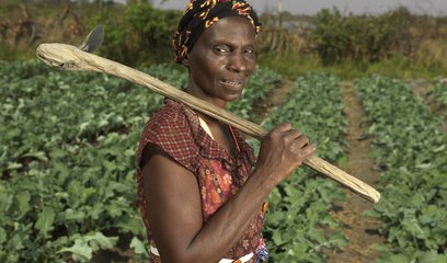 A woman holds a wooden farming implement standing confidently in front of a field of crops