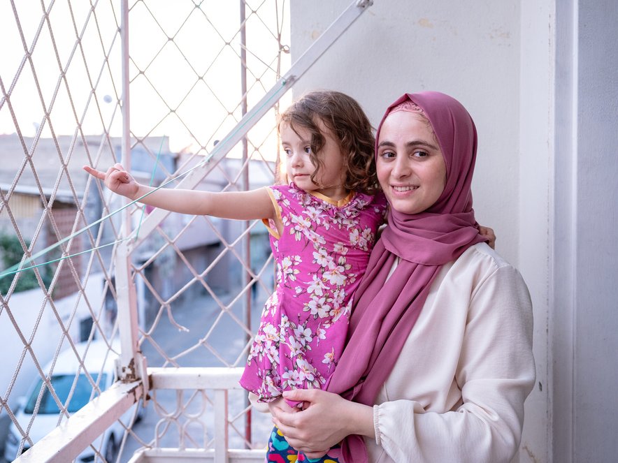 A young woman holds a young girl in her arms on a balcony. The girl is reaching out towards a wire grille that covers the balcony.