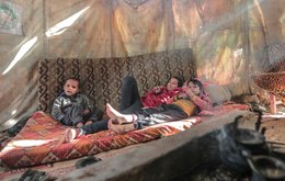 Three children sit on a mattress inside a makeshift tent