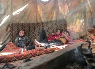 Three children sit on a mattress inside a makeshift tent