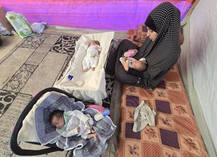 A woman in a tent feeds a baby with a bottle in her lap. There is another baby in a cot in front of her, and one in a car seat next to her