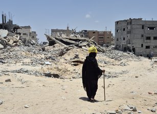 A person dressed all in black walks with a stick amid the rubble of destroyed buildings