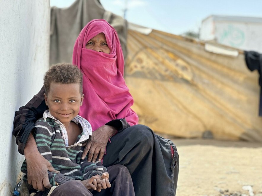 A woman with her arm around a young boy; a refugee camp is visible in the background
