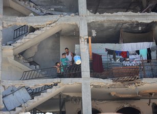 A man and two young children on the balcony of a damaged building whose outer walls have been ripped away by bombing