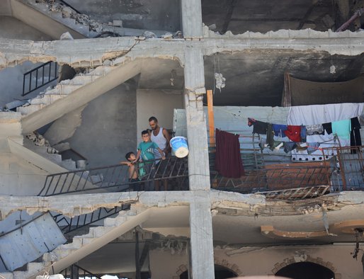 A man and two young children on the balcony of a damaged building whose outer walls have been ripped away by bombing