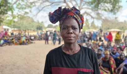 A woman wearing a headscarf stands in the foreground, in front of a large group of people sitting and standing together at a camp for displaced people