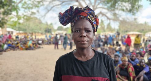 A woman wearing a headscarf stands in the foreground, in front of a large group of people sitting and standing together at a camp for displaced people