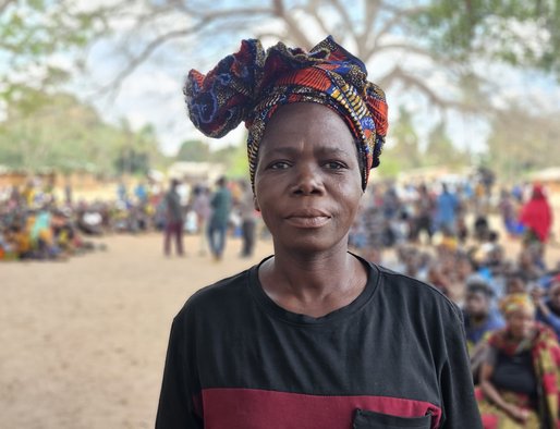 A woman wearing a headscarf stands in the foreground, in front of a large group of people sitting and standing together at a camp for displaced people