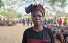 A woman wearing a headscarf stands in the foreground, in front of a large group of people sitting and standing together at a camp for displaced people