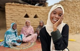 A young woman stands in the foreground, smiling with one hand to her face. three women are sitting on the floor behind her.