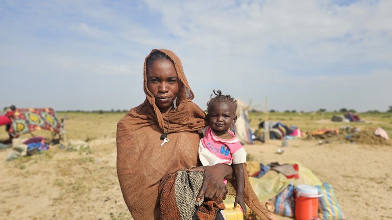 A woman holding a baby. In the background are groups of refugees in an arid landscape.