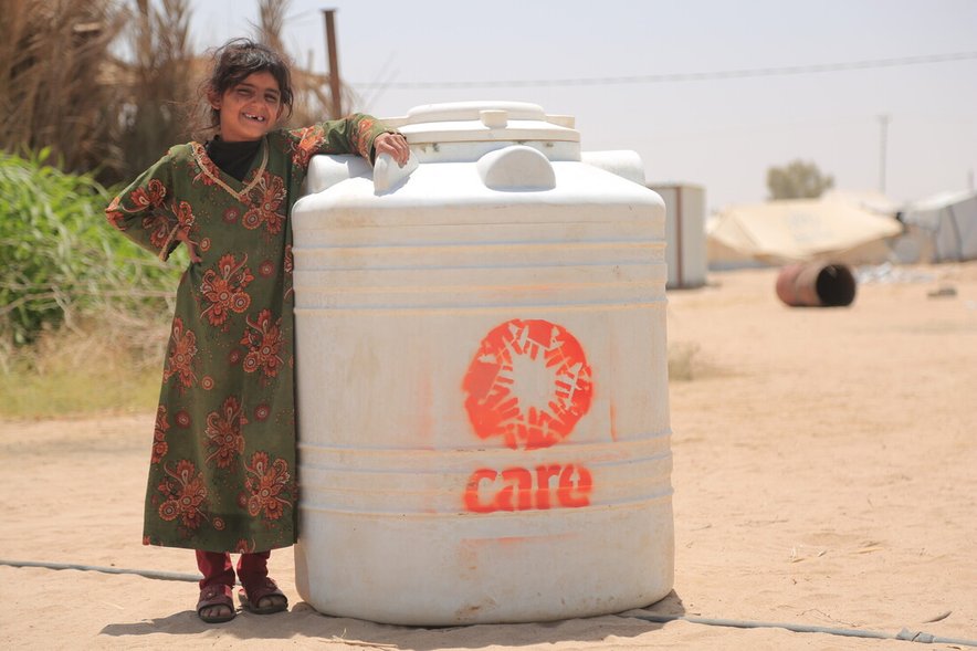 A girl stands next to a large water butt with a CARE log on it