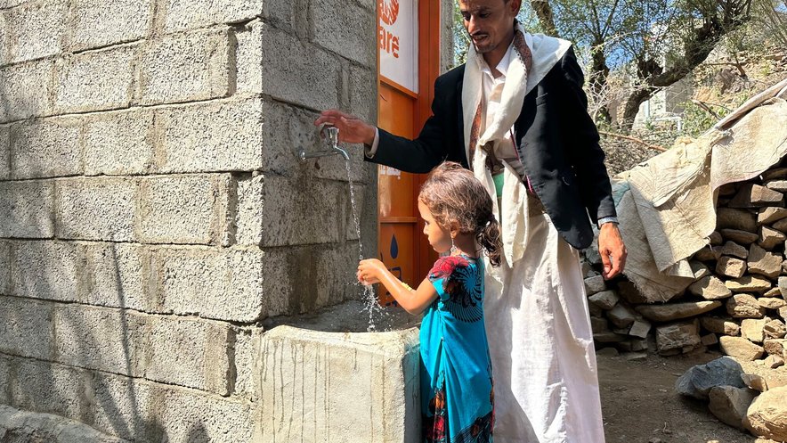 A young girl washes her hands in a stone water fountain with a CARE log on oit. A man is standing with her.