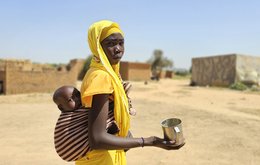 A woman in an arid landscape holds a baby in a sling on her back, and an empty mug in her hand