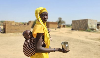 A woman in an arid landscape holds a baby in a sling on her back, and an empty mug in her hand