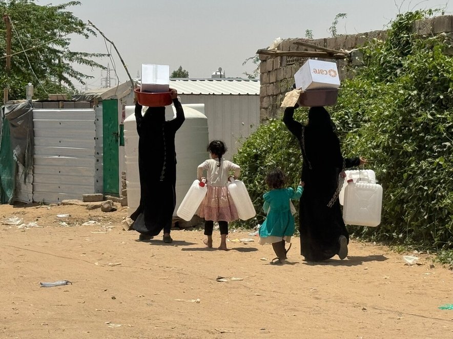 Two women and two young children walk along a dusty road, the women carry boxes with the CARE logo and one of the girls carries two water containers