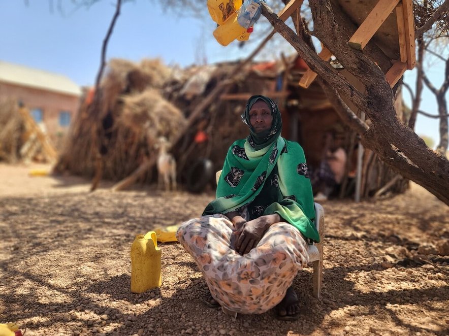 A woman sits in the shade of a tree in a very dry landscape. A yellow water container is next to her, and a wooden hut is visible in the background
