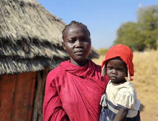 a woman holds a young child standing in front of a hut